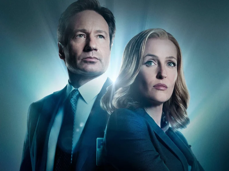 Deux personnes en tenue de travail se tiennent dos à dos avec des expressions sérieuses, éclairées par une lumière bleue provenant de l'arrière, évoquant l'atmosphère mystérieuse de The X-files et rappelant les rôles emblématiques de Gillian Anderson.