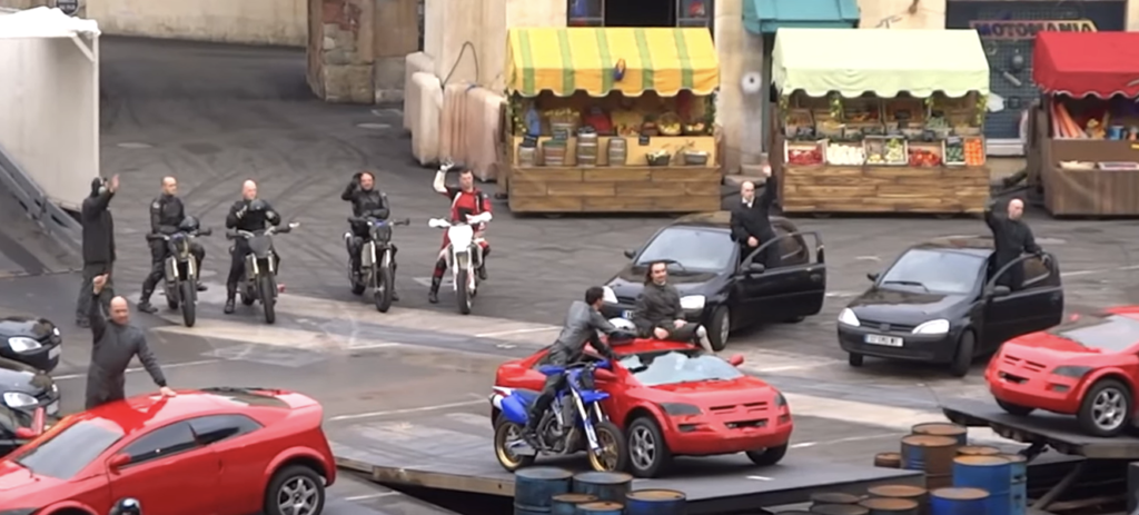 Un groupe de personnes sur des motos et dans des voitures sont positionnées devant des étals de marché, faisant des signes de la main lors d'une mise en scène Moteurs Action Stunt Show Spectacular street scene with red sports cars and barrels avant sa fermeture le 13 mars 2020.