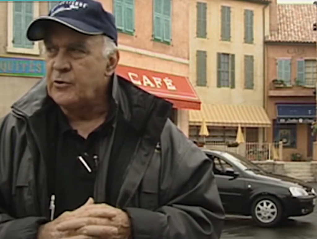 Un homme âgé, vêtu d'une veste sombre et d'une casquette bleue, se tient devant un café à l'auvent rouge, comme s'il se souvenait de la fermeture du 13 mars 2020 ; une voiture noire est garée dans la rue derrière lui.