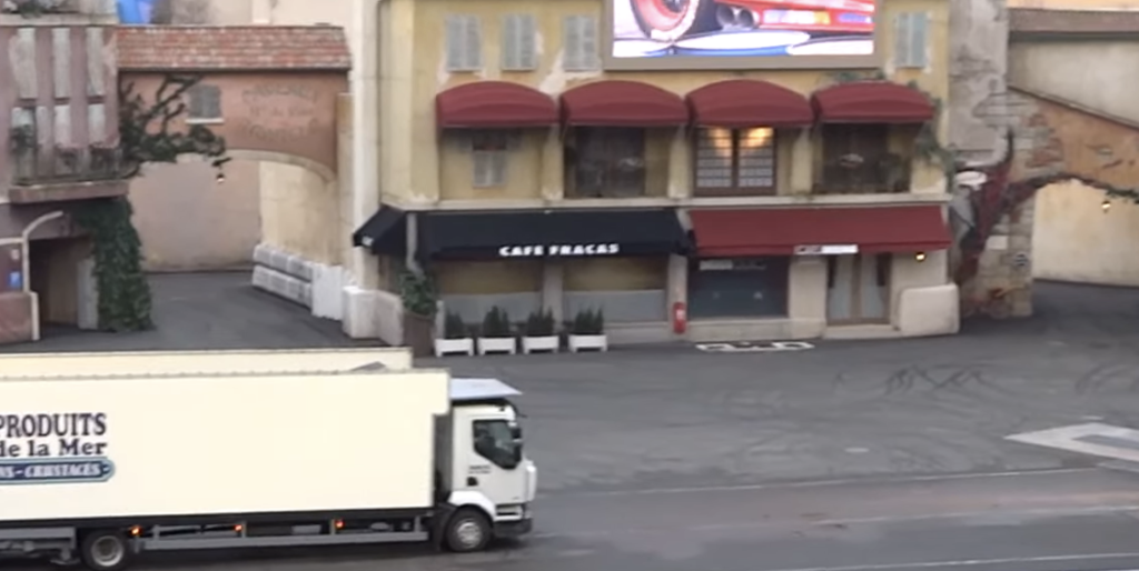 Un camion blanc passe devant un bâtiment jaune avec des auvents rouges et une enseigne "Café Fracas", près de l'ancien Moteurs... Action ! Stunt Show Spectacular, qui a vu sa fermeture le 13 mars 2020, dans une zone pavée vide.