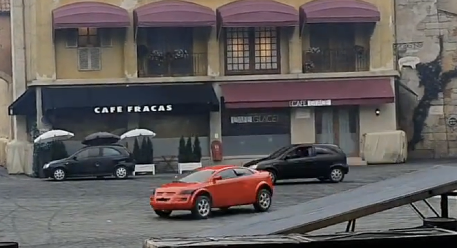 Deux voitures, l'une rouge et l'autre noire, passent à toute vitesse devant deux cafés situés sur une place de la ville avec des parasols et des sièges en plein air - ce qui suscite l'excitation des Moteurs... Action ! Stunt Show Spectacular avant sa fermeture le 13 mars 2020.