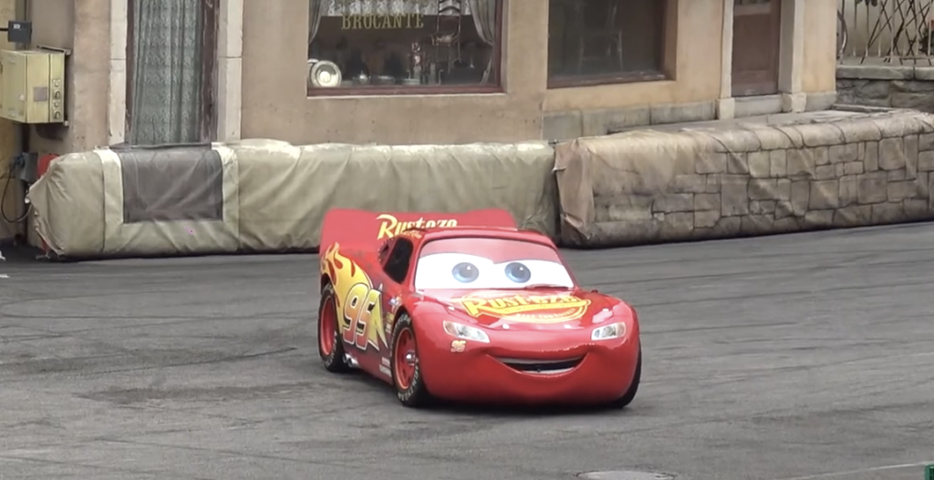 Une voiture de course rouge animée, avec de grands yeux de dessin animé et un sourire, roule dans une rue pavée devant un mur de pierre et une vitrine, inspirée par l'excitation du Moteurs Action Stunt Show avant sa fermeture le 13 mars 2020.