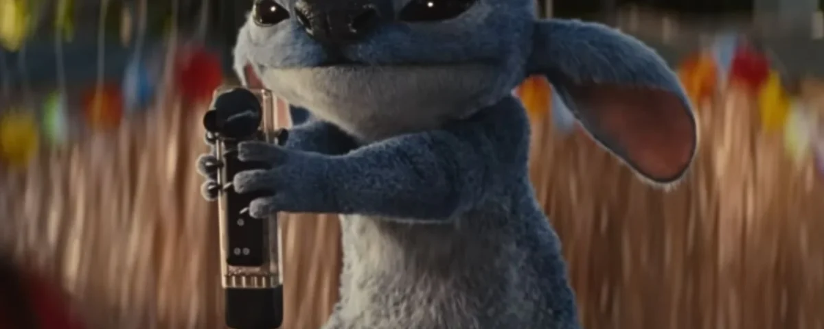 Une créature bleue animée avec de grandes oreilles, inspirée de Lilo & Stitch, tient un microphone tout en se tenant à l'extérieur.