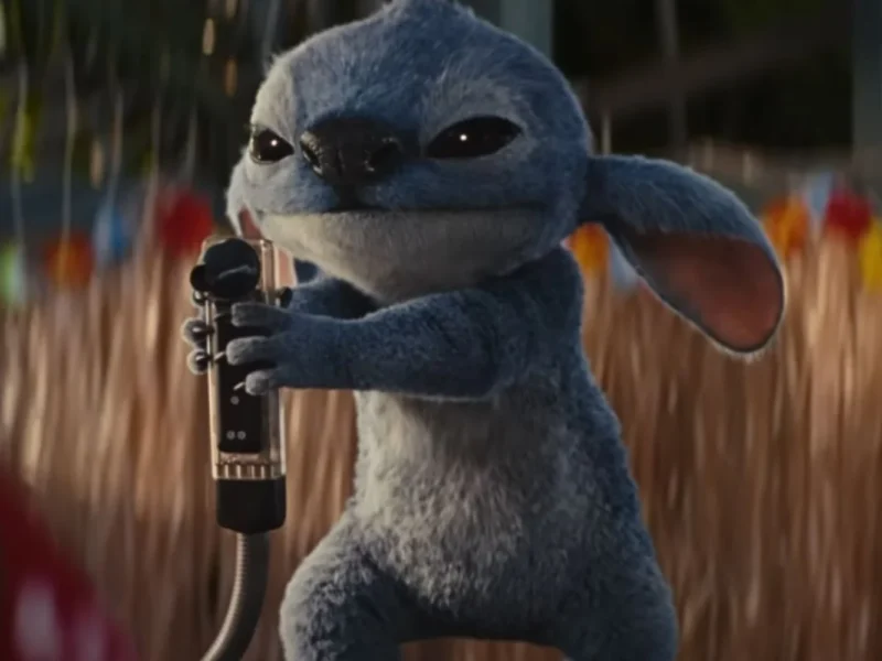 Une créature bleue animée avec de grandes oreilles, inspirée de Lilo & Stitch, tient un microphone tout en se tenant à l'extérieur.