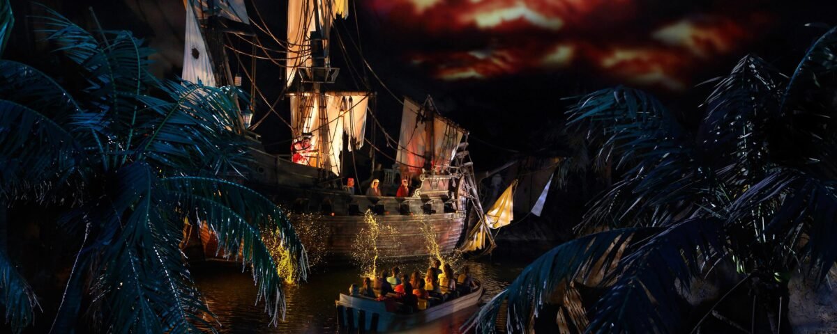 Un bateau de pirates aux voiles en lambeaux fait face à un petit bateau de personnes sur un cours d'eau sombre, entouré de palmiers artificiels sous un ciel rouge dramatique - l'une des attractions à sensations de Disneyland Paris.