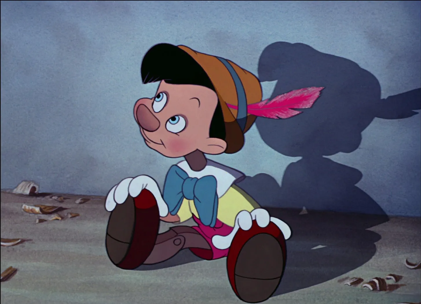 La marionnette animée en bois, coiffée d'un chapeau à plumes, est assise sur le sol et regarde vers le haut, projetant une ombre de forme humaine - un clin d'œil à l'influence culturelle que l'on retrouve dans les films classiques de Disney.