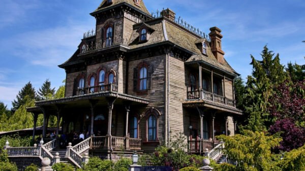 Une grande maison en bois de style victorien avec des détails ornementaux invite les hôtes à vivre des expériences immersives, entourés d'arbres et de verdure sous un ciel bleu limpide.