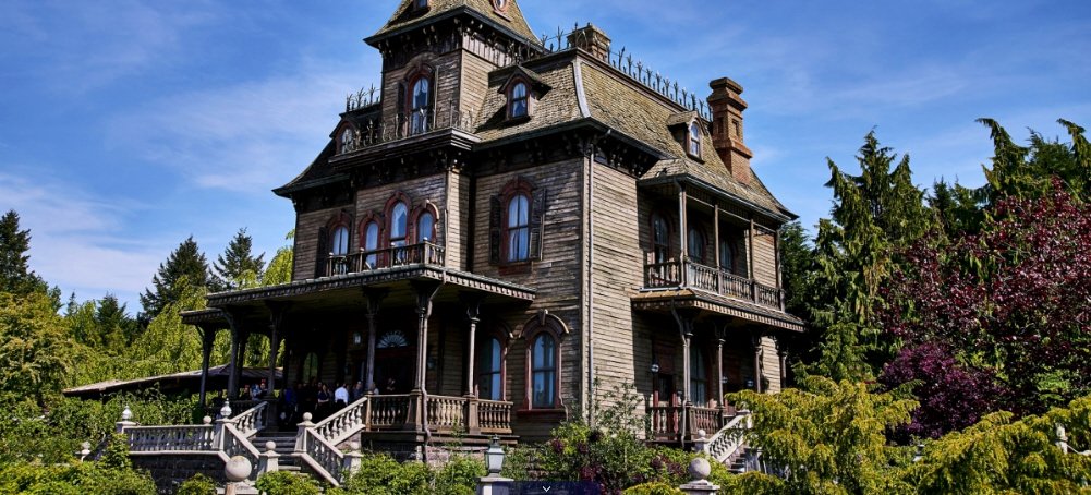 Une grande maison en bois de style victorien avec des détails ornementaux invite les hôtes à vivre des expériences immersives, entourés d'arbres et de verdure sous un ciel bleu limpide.