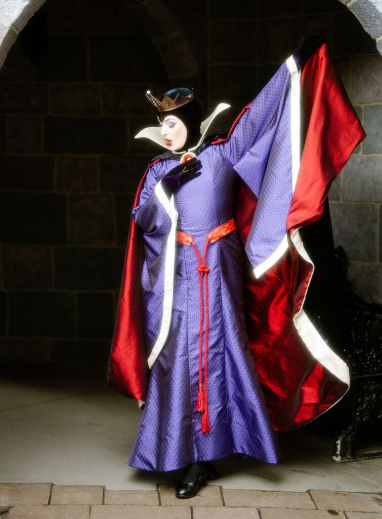 Une personne vêtue d'une robe violette, d'une cape bordée de rouge, d'un col haut blanc et d'une couronne prend une pose dramatique contre un mur de pierre, incarnant Sabrina Von B. dans une scène digne de devenir une vidéo virale.