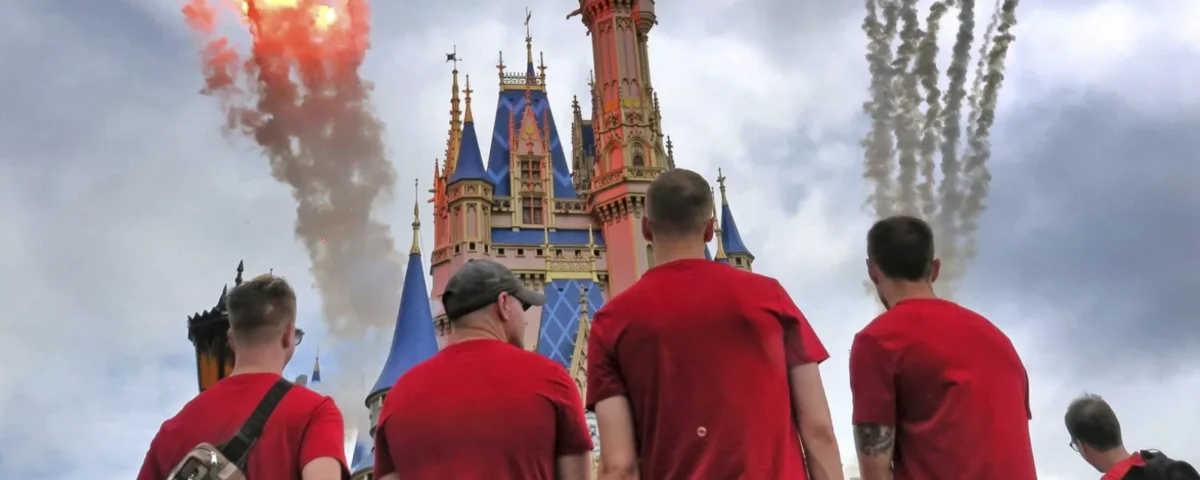 Quatre personnes en chemise rouge se tiennent face à un château coloré à Walt Disney World, alors que des feux d'artifice et de la fumée explosent dans le ciel nuageux, célébrant les Gay Days 2026 avec leur communauté.
