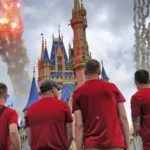 Quatre personnes en chemise rouge se tiennent face à un château coloré à Walt Disney World, alors que des feux d'artifice et de la fumée explosent dans le ciel nuageux, célébrant les Gay Days 2026 avec leur communauté.