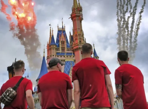 Quatre personnes en chemise rouge se tiennent face à un château coloré à Walt Disney World, alors que des feux d'artifice et de la fumée explosent dans le ciel nuageux, célébrant les Gay Days 2026 avec leur communauté.