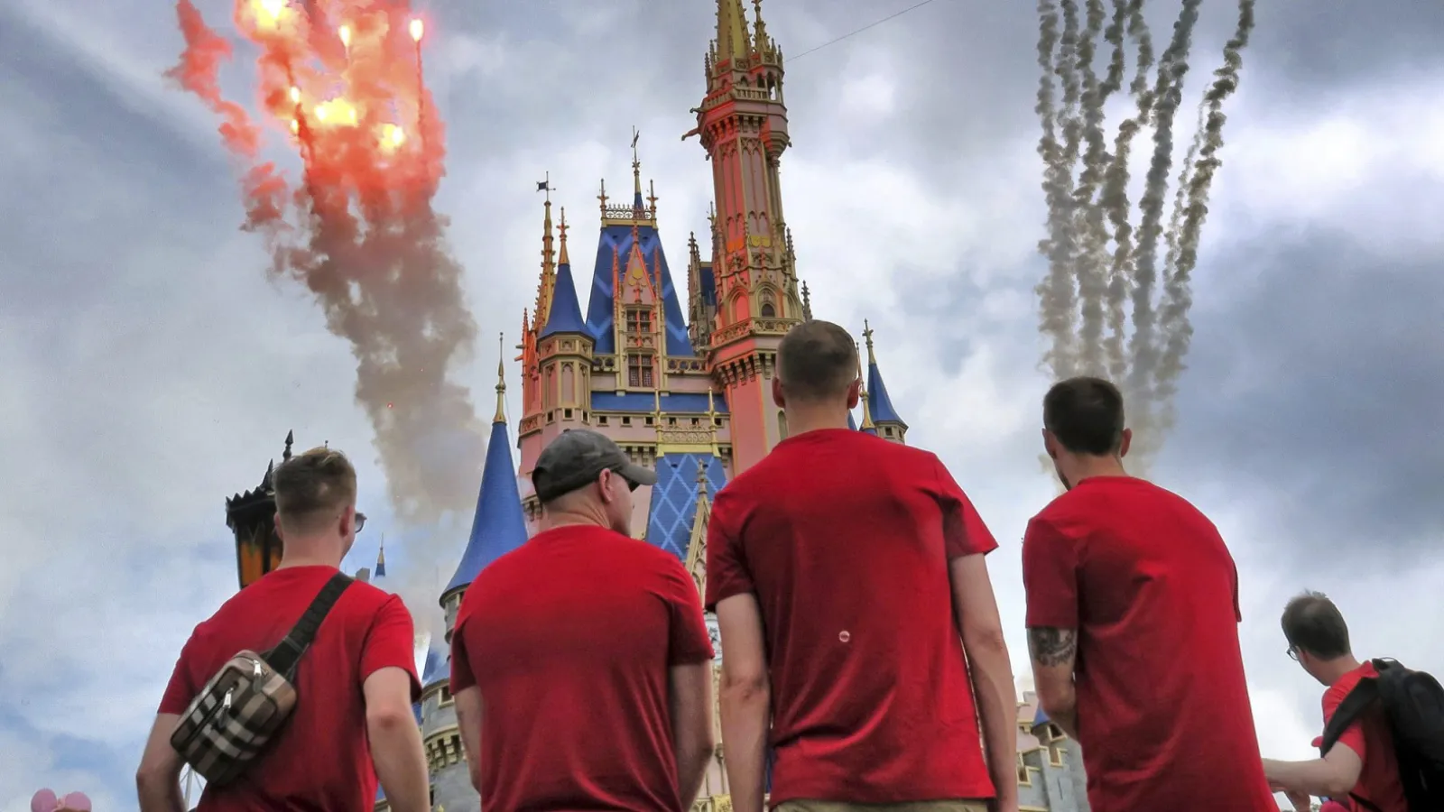 Quatre personnes en chemise rouge se tiennent face à un château coloré à Walt Disney World, alors que des feux d'artifice et de la fumée explosent dans le ciel nuageux, célébrant les Gay Days 2026 avec leur communauté.