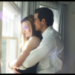 Un homme vêtu d'une chemise blanche se tient derrière une femme vêtue d'un haut sans manches, l'enlaçant alors qu'ils regardent par une fenêtre ensoleillée - évoquant l'élégance intemporelle de John F. Kennedy Jr. et de Carolyn Bessette dans une pièce doucement éclairée.