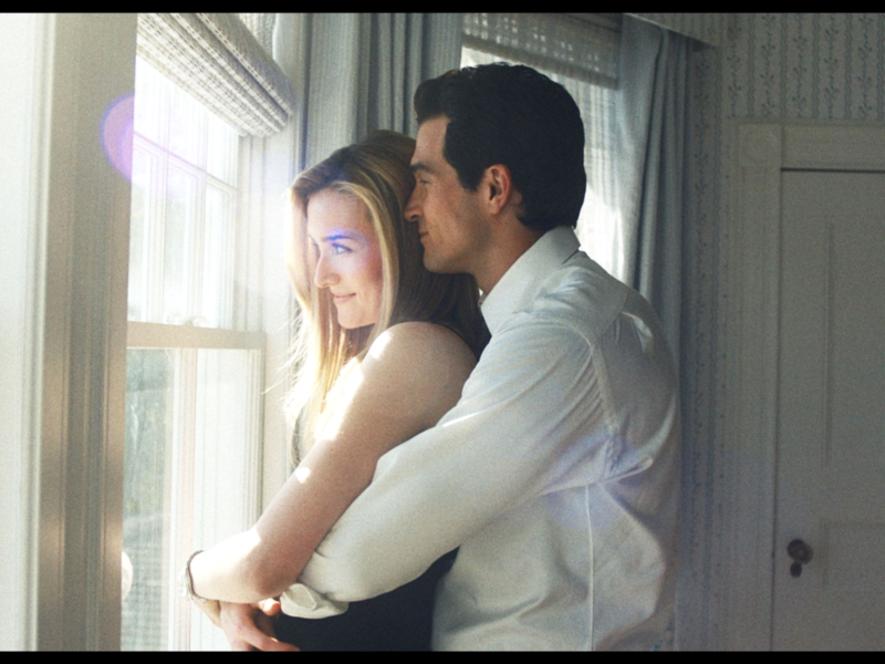 Un homme vêtu d'une chemise blanche se tient derrière une femme vêtue d'un haut sans manches, l'enlaçant alors qu'ils regardent par une fenêtre ensoleillée - évoquant l'élégance intemporelle de John F. Kennedy Jr. et de Carolyn Bessette dans une pièce doucement éclairée.