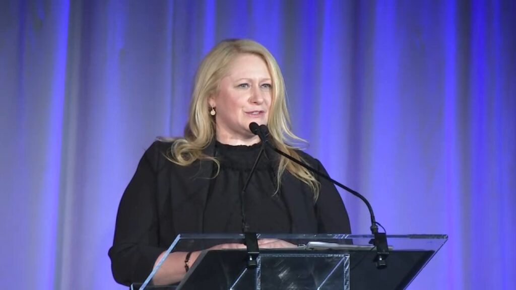 Debra Oconnell, une femme aux longs cheveux blonds, s'exprime sur un podium clair muni d'un microphone, devant un rideau bleu en toile de fond, lors d'un événement organisé par Disney Entertainment Television.