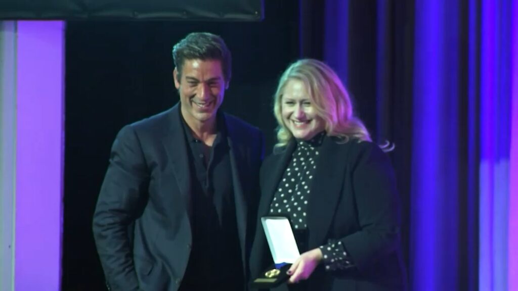 Deux personnes en tenue de soirée se tiennent ensemble sur scène et sourient à la caméra. L'une d'elles tient une médaille dans une boîte ouverte. Des lumières bleues et violettes illuminent l'arrière-plan, célébrant un moment spécial à Disney Entertainment Television.