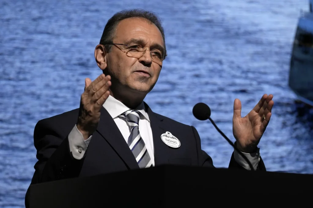 Un homme en costume-cravate, peut-être Thomas Mazloum de Disney's Division Parcs, parle sur un podium avec un microphone, en faisant des gestes des deux mains. Un plan d'eau est visible à l'arrière-plan.