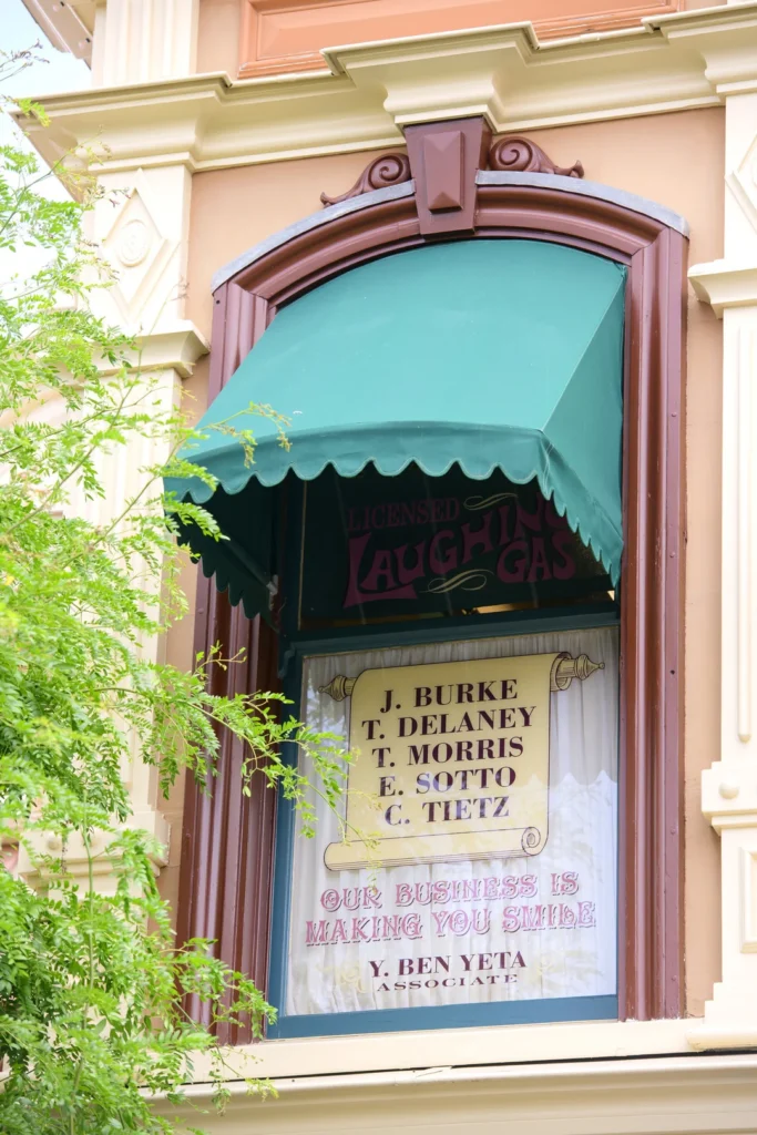 Une fenêtre avec un auvent vert affiche une liste de noms et la phrase "Our business is making you smile" (Notre métier est de vous faire sourire), encadrée par des détails ornés du bâtiment - une touche qui rappelle le travail de l'imagier Eddie Sotto à Disneyland Paris - partiellement masqués par des branches d'arbre.