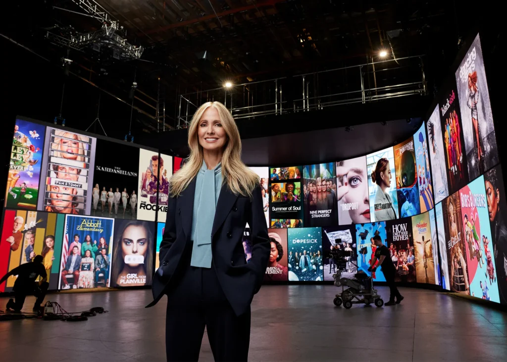 Une femme en tailleur se tient dans un studio entouré de grands écrans affichant diverses affiches d'émissions télévisées et de films, reflétant l'univers dynamique de Disney Entertainment Television.