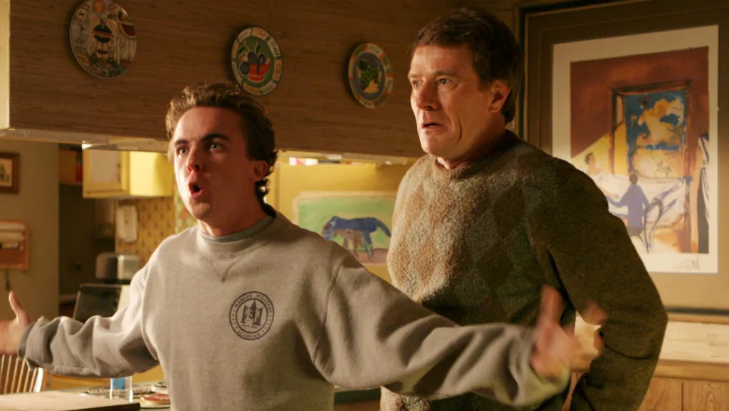 Deux hommes se tiennent dans une cuisine ; le plus jeune, qui ressemble à Malcolm (Frankie Muniz), parle avec emphase, les bras tendus, tandis que le plus âgé, qui ressemble beaucoup au personnage de Bryan Cranston, regarde la scène d'un air inquiet.
