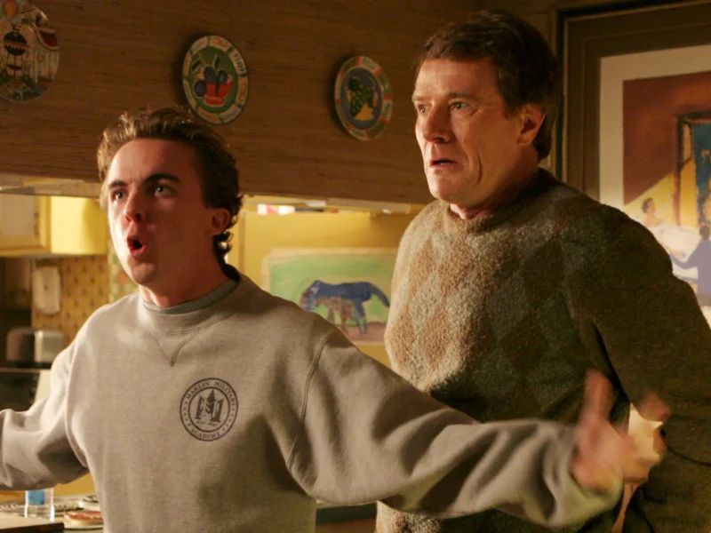 Deux hommes se tiennent dans une cuisine ; le plus jeune, qui ressemble à Malcolm (Frankie Muniz), parle avec emphase, les bras tendus, tandis que le plus âgé, qui ressemble beaucoup au personnage de Bryan Cranston, regarde la scène d'un air inquiet.