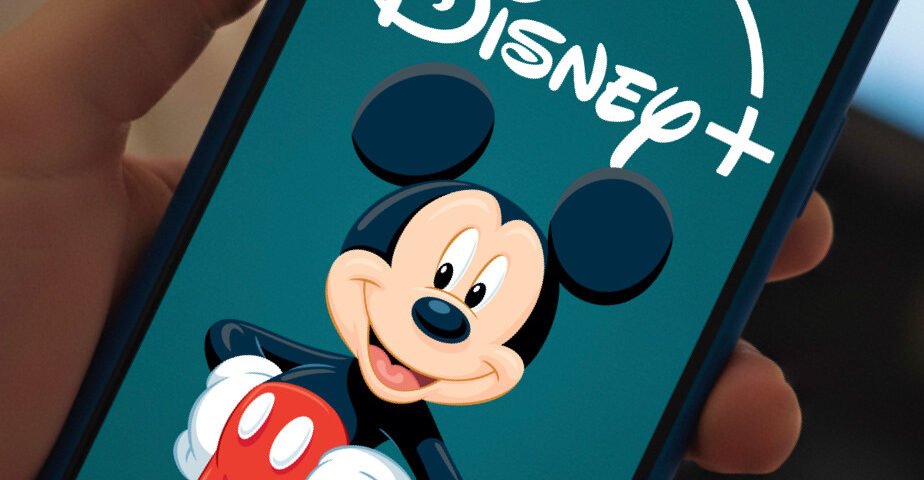 Une main tenant un smartphone affiche le logo Disney+ avec une image de Mickey Mouse sur l'écran, mettant en avant des vidéos verticales conçues pour renforcer l'engagement des abonnés.