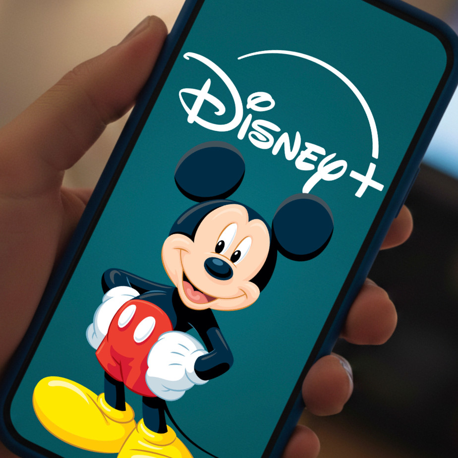 Une main tenant un smartphone affiche le logo Disney+ avec une image de Mickey Mouse sur l'écran, mettant en avant des vidéos verticales conçues pour renforcer l'engagement des abonnés.