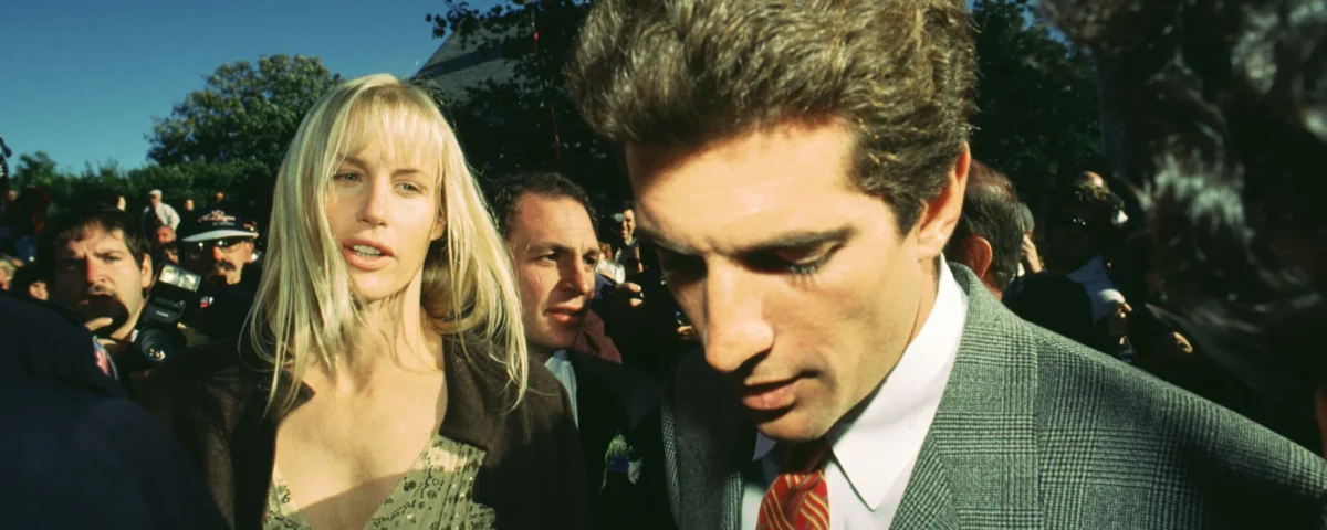 Une femme et un homme en tenue de soirée marchent à l'extérieur dans une foule, avec des arbres et un ciel dégagé en arrière-plan, évoquant l'élégance d'une scène de Love Story Disney+.