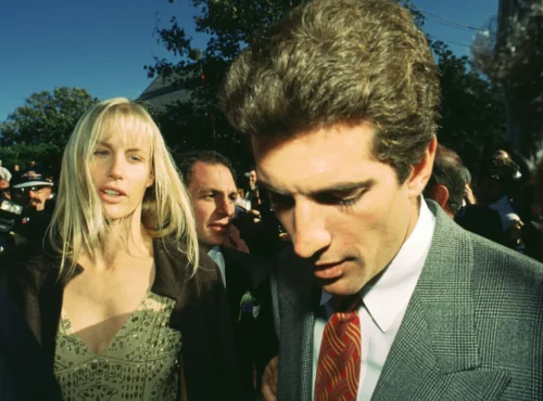 Une femme et un homme en tenue de soirée marchent à l'extérieur dans une foule, avec des arbres et un ciel dégagé en arrière-plan, évoquant l'élégance d'une scène de Love Story Disney+.