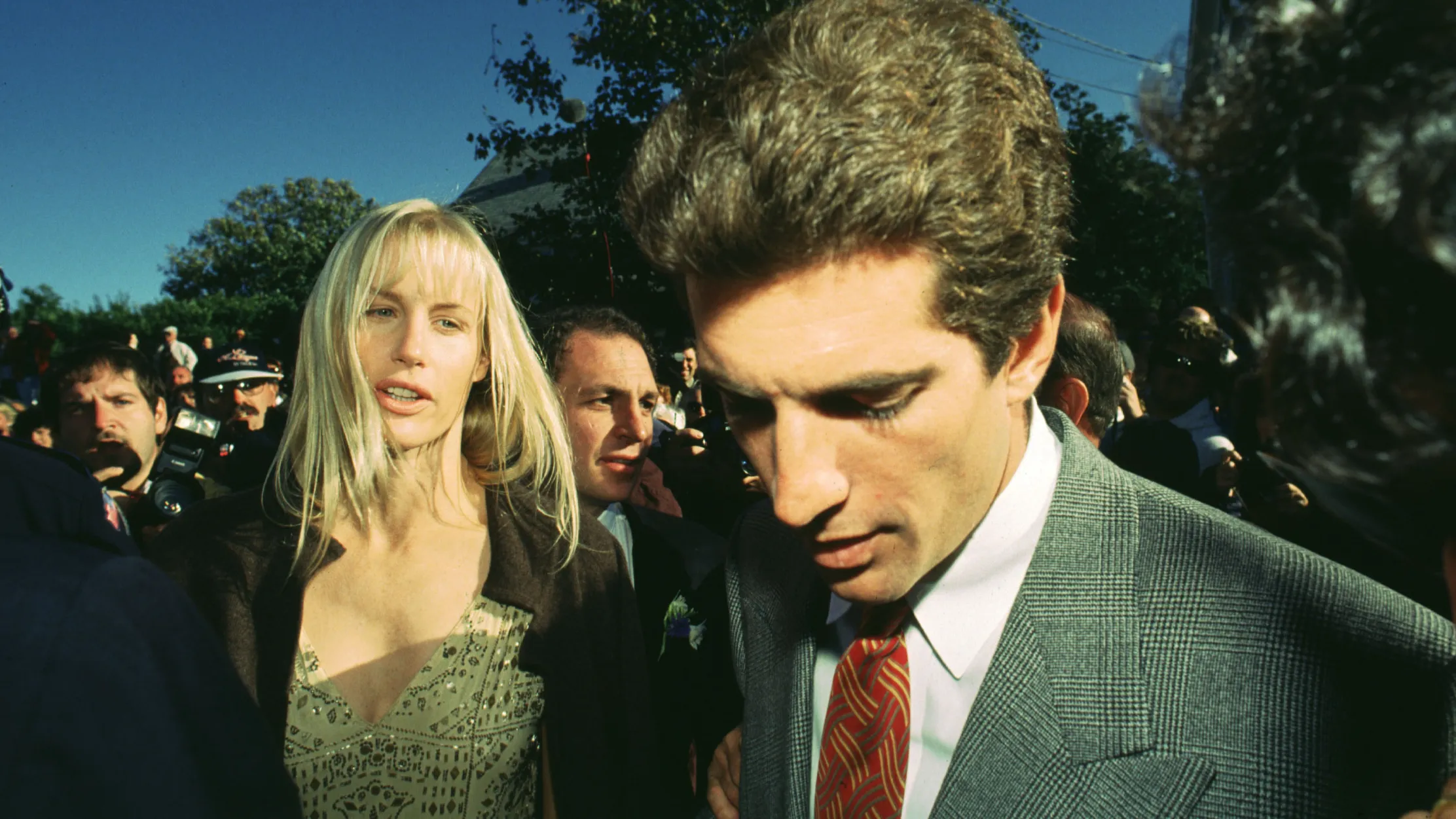 Une femme et un homme en tenue de soirée marchent à l'extérieur dans une foule, avec des arbres et un ciel dégagé en arrière-plan, évoquant l'élégance d'une scène de Love Story Disney+.