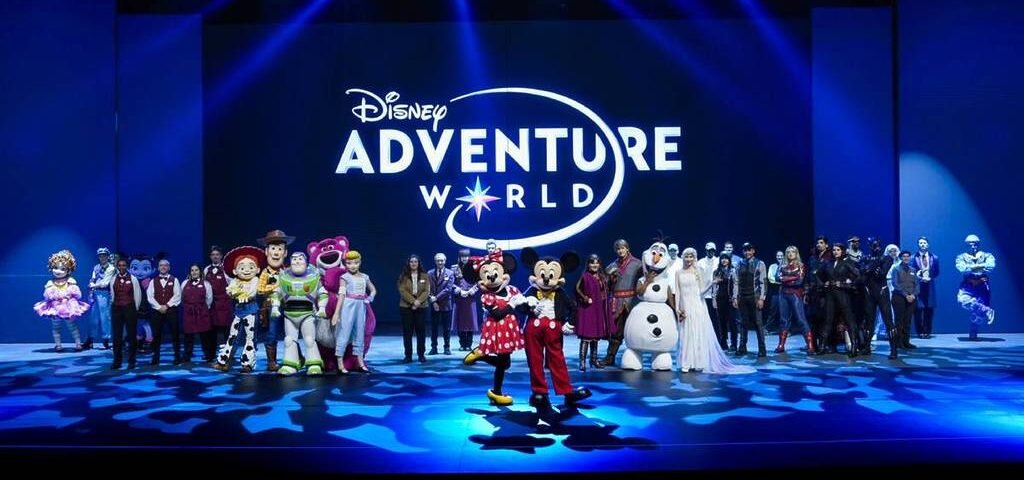 Un groupe de personnages et de personnes de Disney se tient sur scène devant un grand panneau "Disney Adventure World", célébrant la nouvelle campagne à Disneyland Paris.