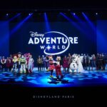 Un groupe de personnages et de personnes de Disney se tient sur scène devant un grand panneau "Disney Adventure World", célébrant la nouvelle campagne à Disneyland Paris.