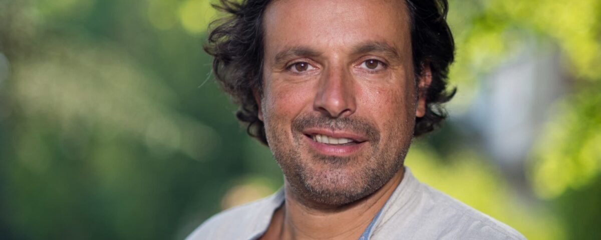 Un homme aux cheveux bruns mi-longs et à la barbe porte une chemise de couleur claire, debout en plein air, avec de la verdure en arrière-plan flou - rappelant Bruno Salomone, connu pour ses rôles dans Disney Doublages Pixar.
