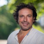 Un homme aux cheveux bruns mi-longs et à la barbe porte une chemise de couleur claire, debout en plein air, avec de la verdure en arrière-plan flou - rappelant Bruno Salomone, connu pour ses rôles dans Disney Doublages Pixar.