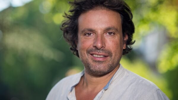 Un homme aux cheveux bruns mi-longs et à la barbe porte une chemise de couleur claire, debout en plein air, avec de la verdure en arrière-plan flou - rappelant Bruno Salomone, connu pour ses rôles dans Disney Doublages Pixar.