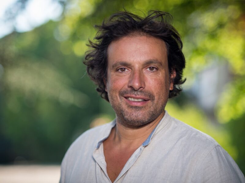 Un homme aux cheveux bruns mi-longs et à la barbe porte une chemise de couleur claire, debout en plein air, avec de la verdure en arrière-plan flou - rappelant Bruno Salomone, connu pour ses rôles dans Disney Doublages Pixar.