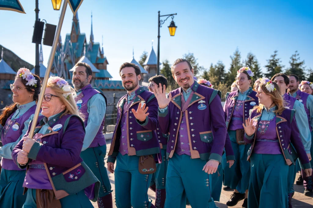 Un groupe de personnes vêtues de tenues coordonnées violettes et sarcelles marchent et saluent en souriant devant un château à Disneyland Paris lors d'un événement en plein air "World Of Frozen".