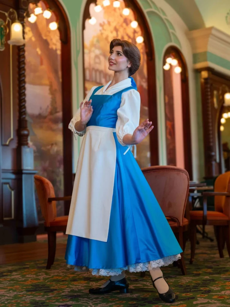 Une personne portant une robe bleue et blanche avec un tablier blanc pose à l'intérieur du restaurant Regal View à Disneyland Paris, entourée d'un décor de style ancien et de chaises rembourrées, rappelant les nouvelles tenues Ariel.