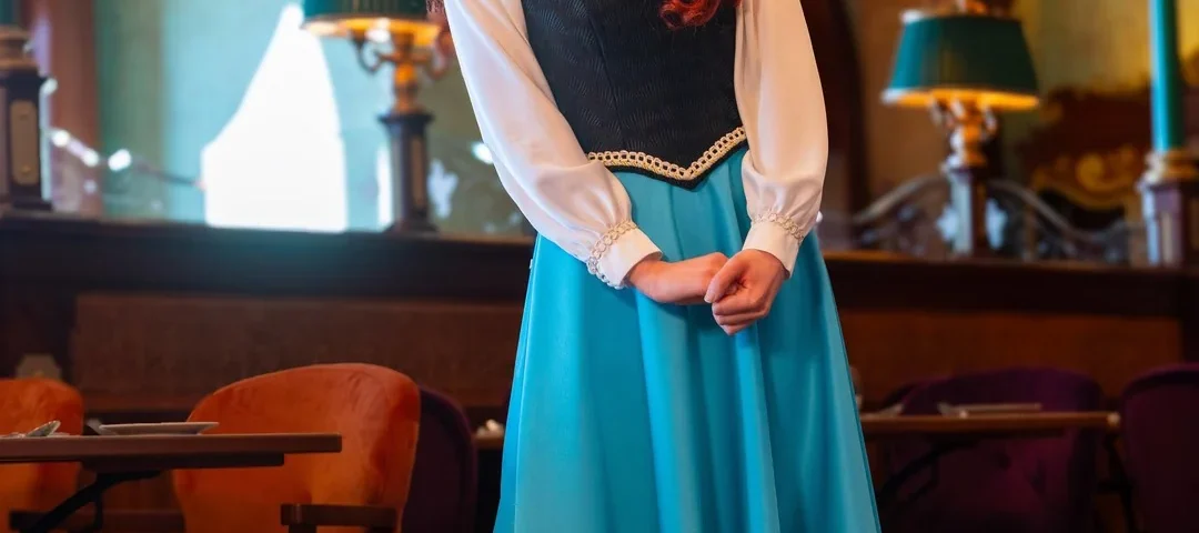 Une femme vêtue d'une jupe bleue, d'un chemisier blanc et d'un corsage noir se tient à l'intérieur du restaurant Regal View, souriante, les mains jointes, entourée de meubles de style ancien et d'un éclairage orné rappelant l'élégance de Disneyland Paris.