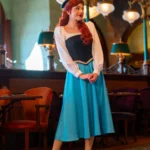 Une femme vêtue d'une jupe bleue, d'un chemisier blanc et d'un corsage noir se tient à l'intérieur du restaurant Regal View, souriante, les mains jointes, entourée de meubles de style ancien et d'un éclairage orné rappelant l'élégance de Disneyland Paris.