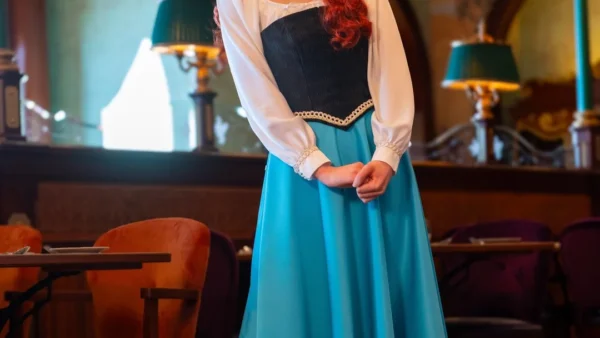 Une femme vêtue d'une jupe bleue, d'un chemisier blanc et d'un corsage noir se tient à l'intérieur du restaurant Regal View, souriante, les mains jointes, entourée de meubles de style ancien et d'un éclairage orné rappelant l'élégance de Disneyland Paris.
