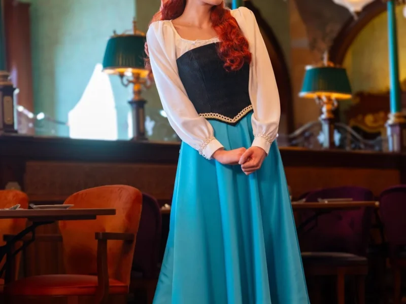 Une femme vêtue d'une jupe bleue, d'un chemisier blanc et d'un corsage noir se tient à l'intérieur du restaurant Regal View, souriante, les mains jointes, entourée de meubles de style ancien et d'un éclairage orné rappelant l'élégance de Disneyland Paris.