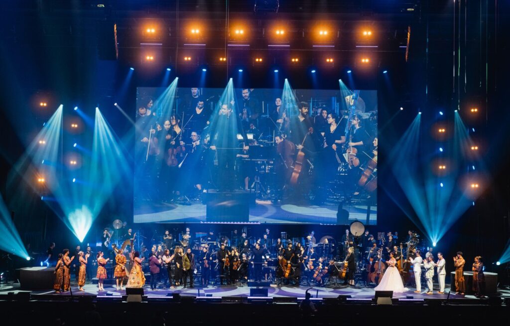 Un grand orchestre et des artistes se tiennent sur scène sous des lumières vives, leur image étant projetée sur un grand écran derrière eux pendant le ciné-concert symphonique Disney : Disney En Concert, dans le cadre de la tournée France 2026.
