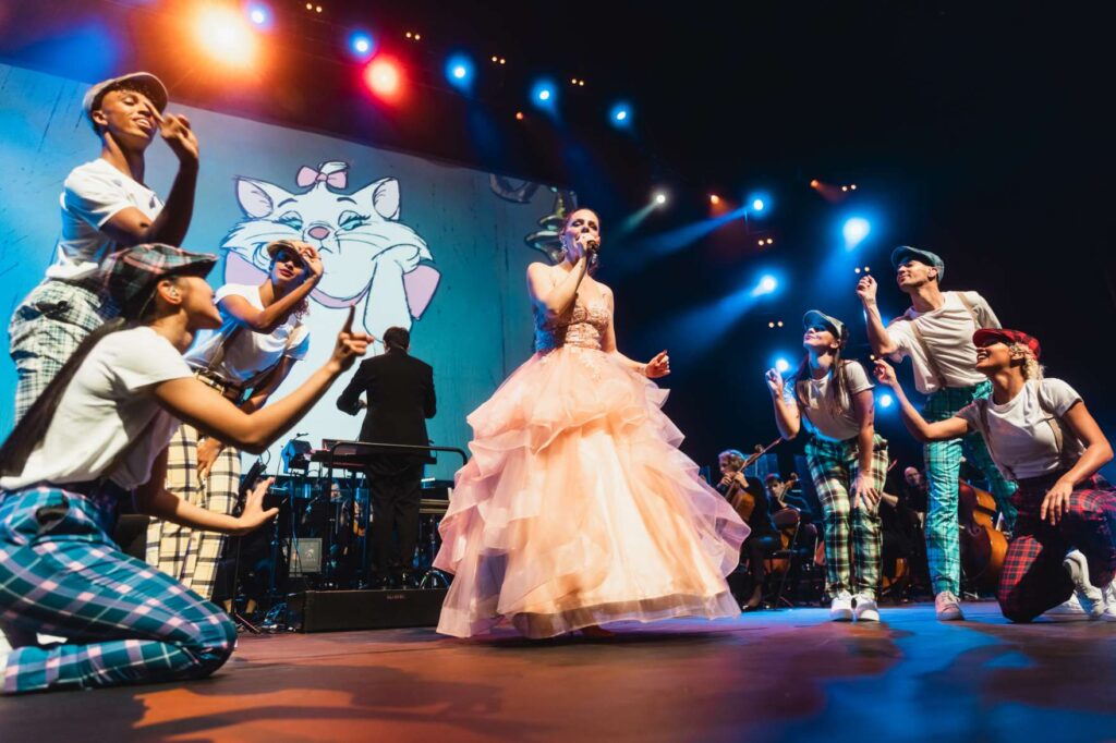 Une femme en robe de bal pêche chante sur scène pendant Disney en concert, entourée de danseurs en pantalon écossais et chemise blanche, avec un orchestre et une image de chat animé en arrière-plan, dans le cadre du ciné-concert symphonique tournée France.