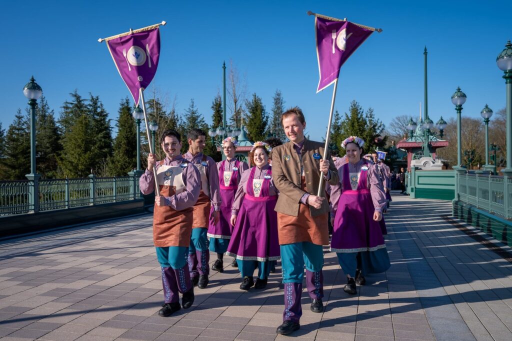 Un groupe de personnes vêtues de costumes coordonnés violets et sarcelles marche en plein air sur un chemin pavé en formation précise, deux d'entre elles tenant des drapeaux violets, avec des arbres et des lampadaires en arrière-plan.