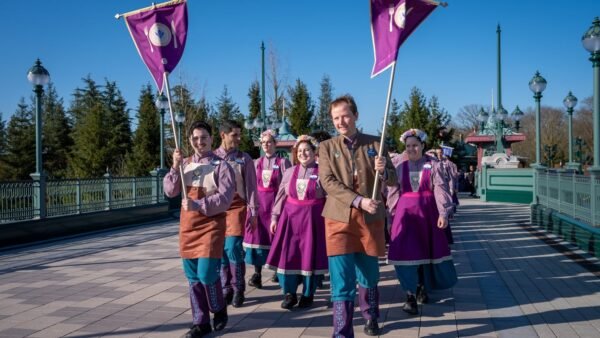 Un groupe de personnes vêtues de costumes coordonnés violets et sarcelles marche en plein air sur un chemin pavé en formation précise, deux d'entre elles tenant des drapeaux violets, avec des arbres et des lampadaires en arrière-plan.