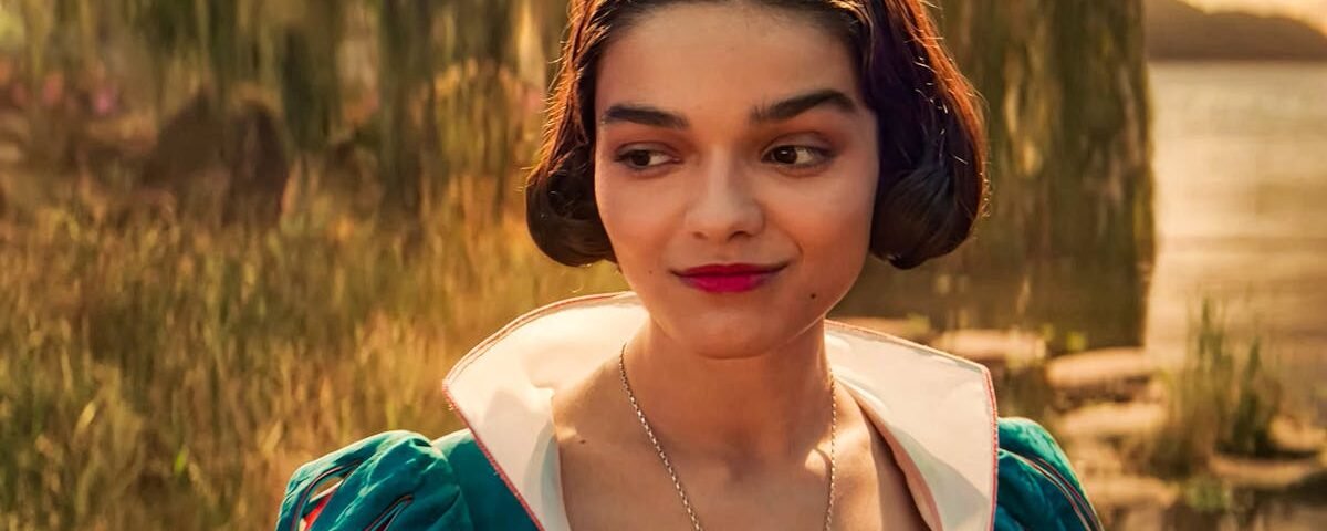 Une femme vêtue d'une robe sarcelle aux manches bouffantes se tient à l'extérieur, près de l'eau et de la verdure, regardant sur le côté avec un léger sourire, rappelant la gracieuse interprétation de Rachel Zegler dans Blanche-neige.