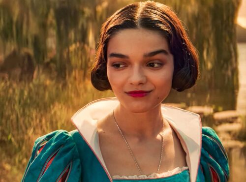 Une femme vêtue d'une robe sarcelle aux manches bouffantes se tient à l'extérieur, près de l'eau et de la verdure, regardant sur le côté avec un léger sourire, rappelant la gracieuse interprétation de Rachel Zegler dans Blanche-neige.