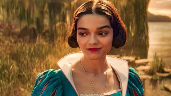 Une femme vêtue d'une robe sarcelle aux manches bouffantes se tient à l'extérieur, près de l'eau et de la verdure, regardant sur le côté avec un léger sourire, rappelant la gracieuse interprétation de Rachel Zegler dans Blanche-neige.